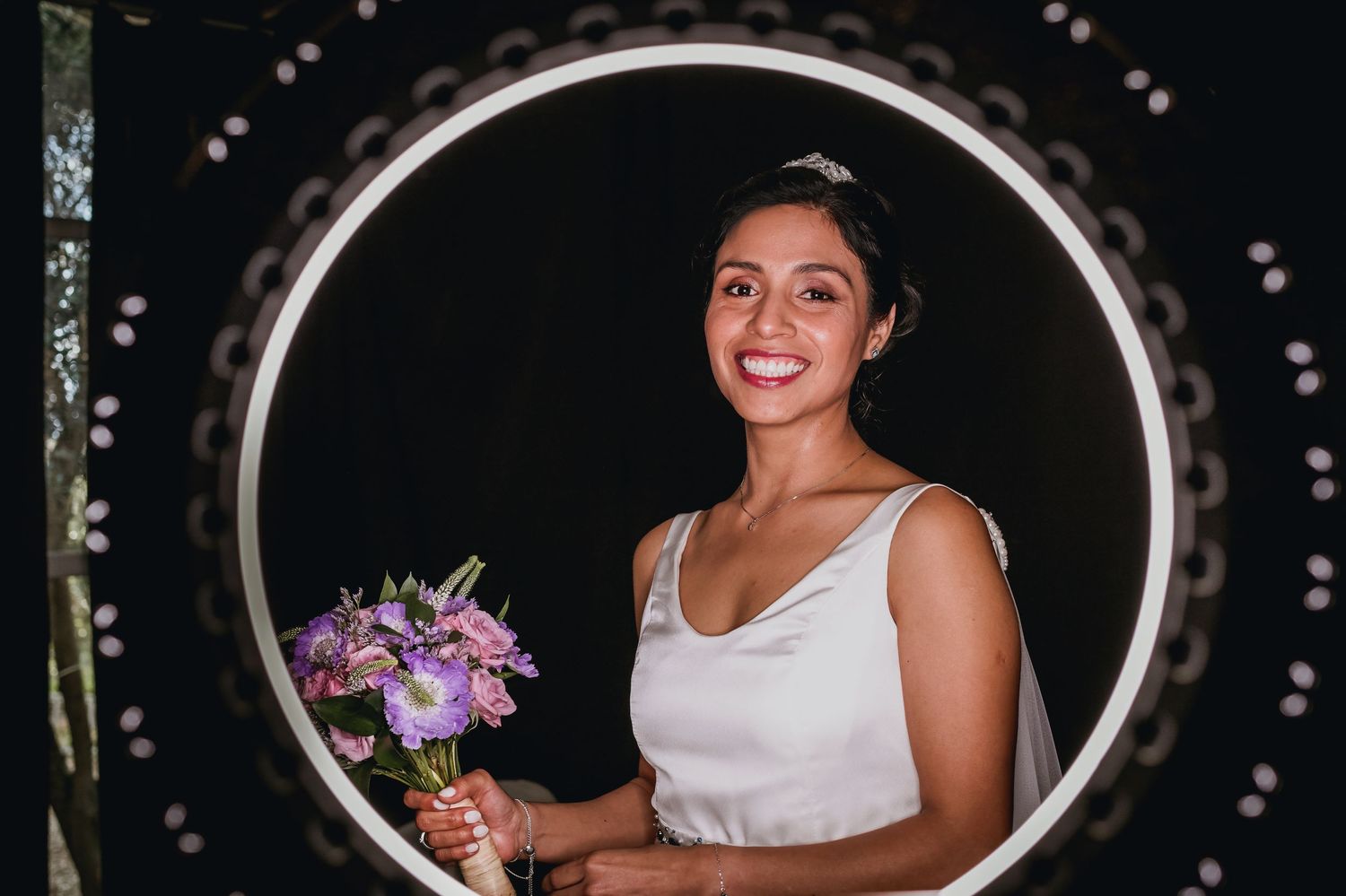 Smiling bride holding purple bouquet posed in illuminated circular frame against dark background.