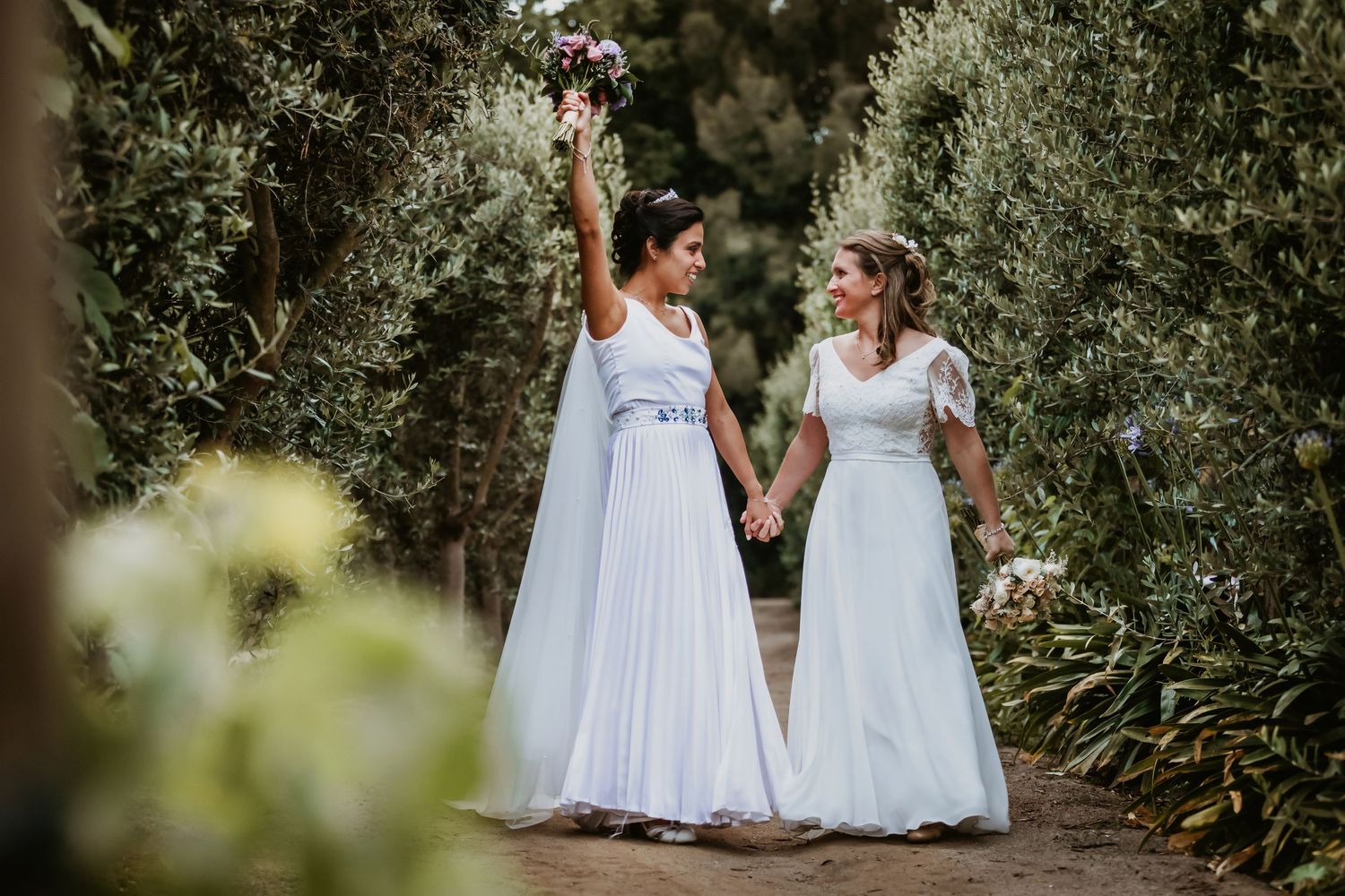 Two brides hold hands and celebrate walking through a tree-lined path on their wedding day.