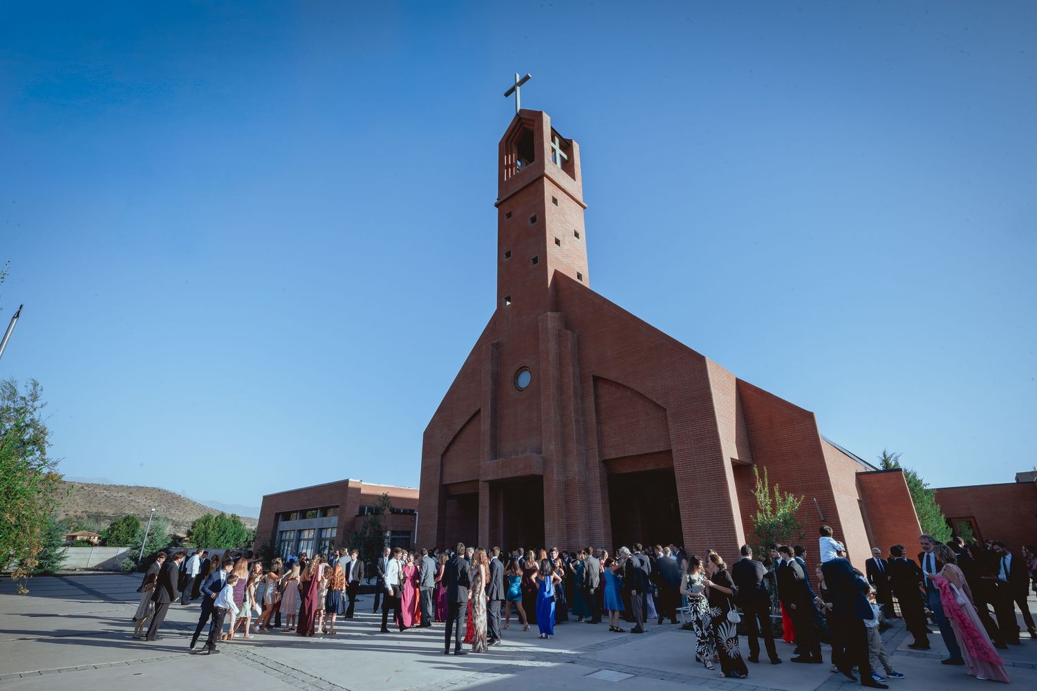 Guests gather outside red brick church with bell tower under clear blue sky.