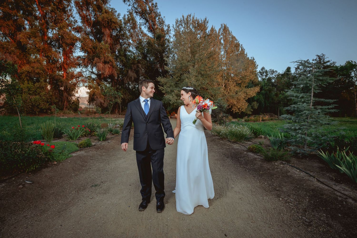 Bride and groom walking hand in hand along a garden path surrounded by autumn foliage.