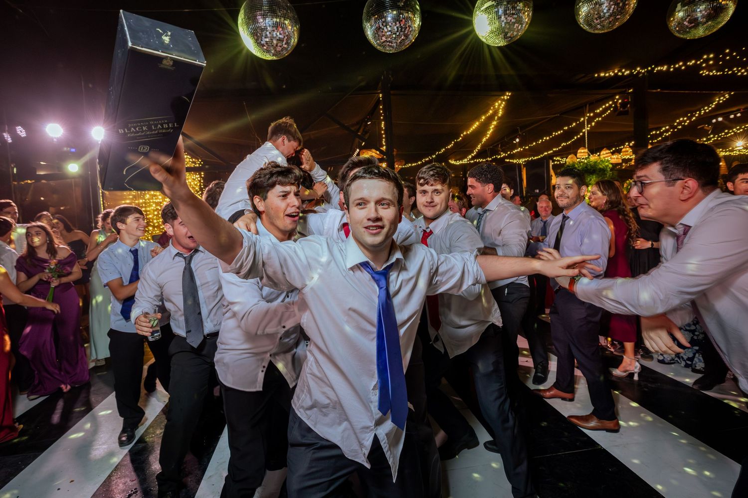 Groomsmen dance energetically on white dance floor at wedding reception with disco balls.