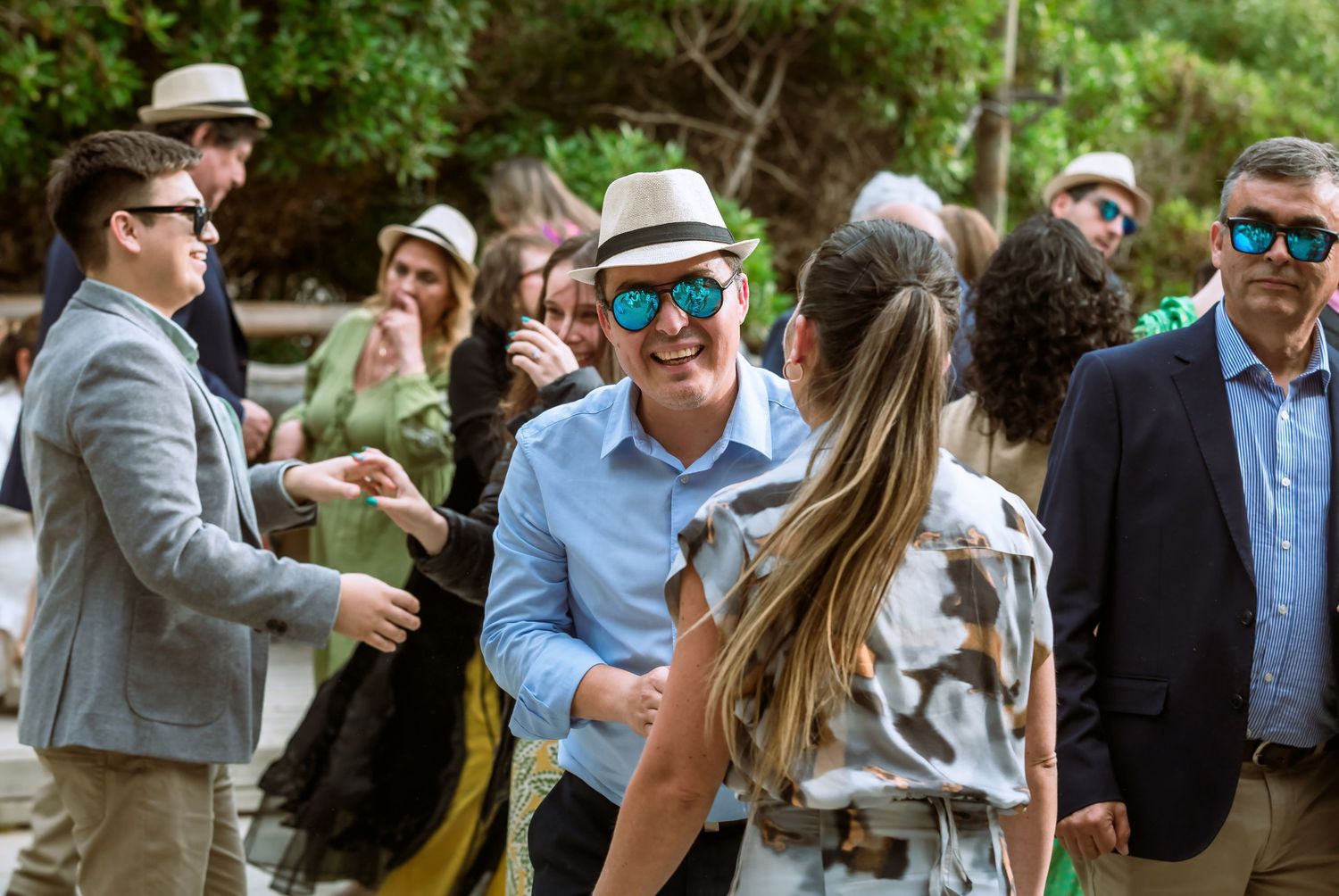 Wedding guests laughing and socializing outdoors, man in panama hat and mirrored sunglasses.