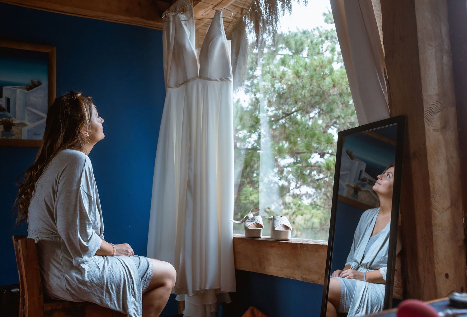 Bride in a robe admires her wedding dress hanging by a window, reflected in a mirror.