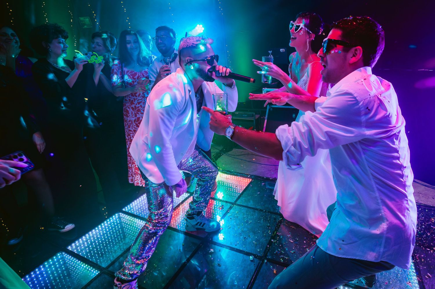 Two performers in white shirts and silver pants energetically perform on a colorful neon-lit stage.