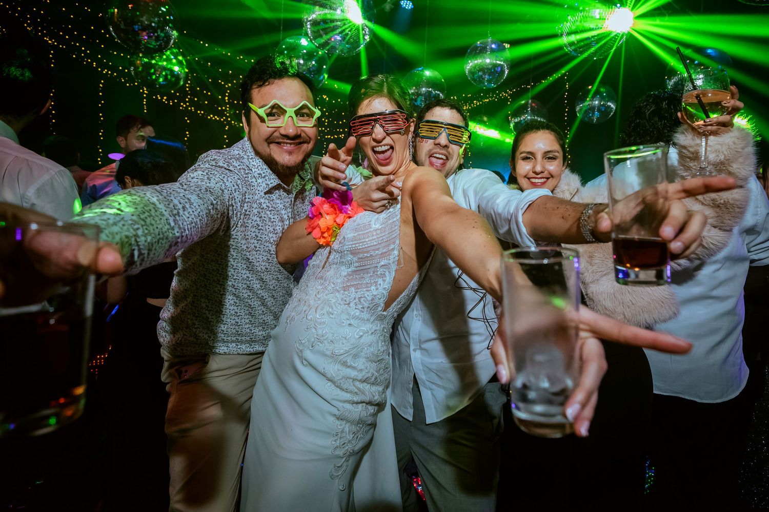 Wedding couple and friends celebrating on dance floor with neon lights, fun glasses, and drinks at a lively reception party.
