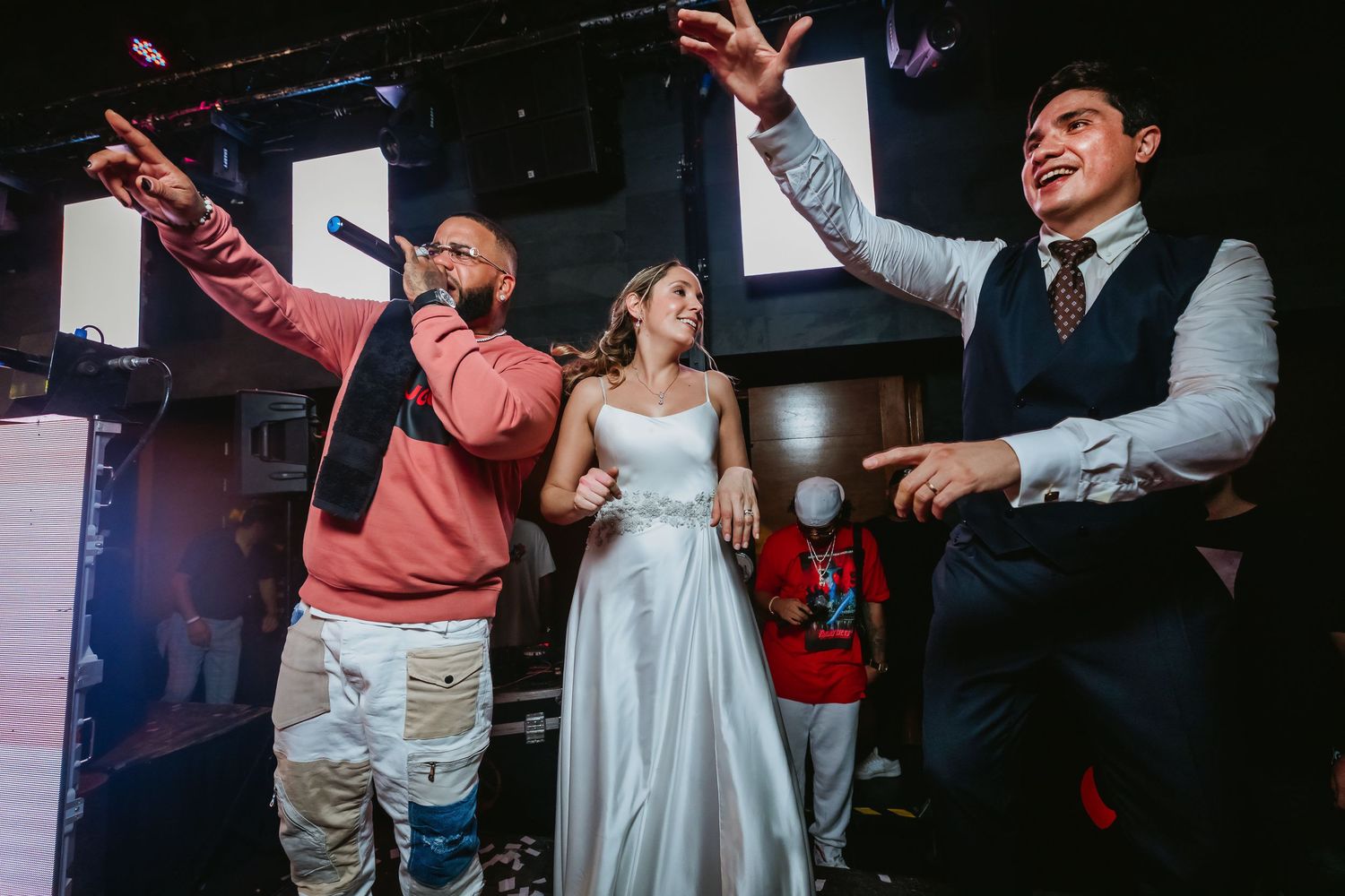 Performer with microphone celebrating on stage with bride in white gown and groom at wedding.