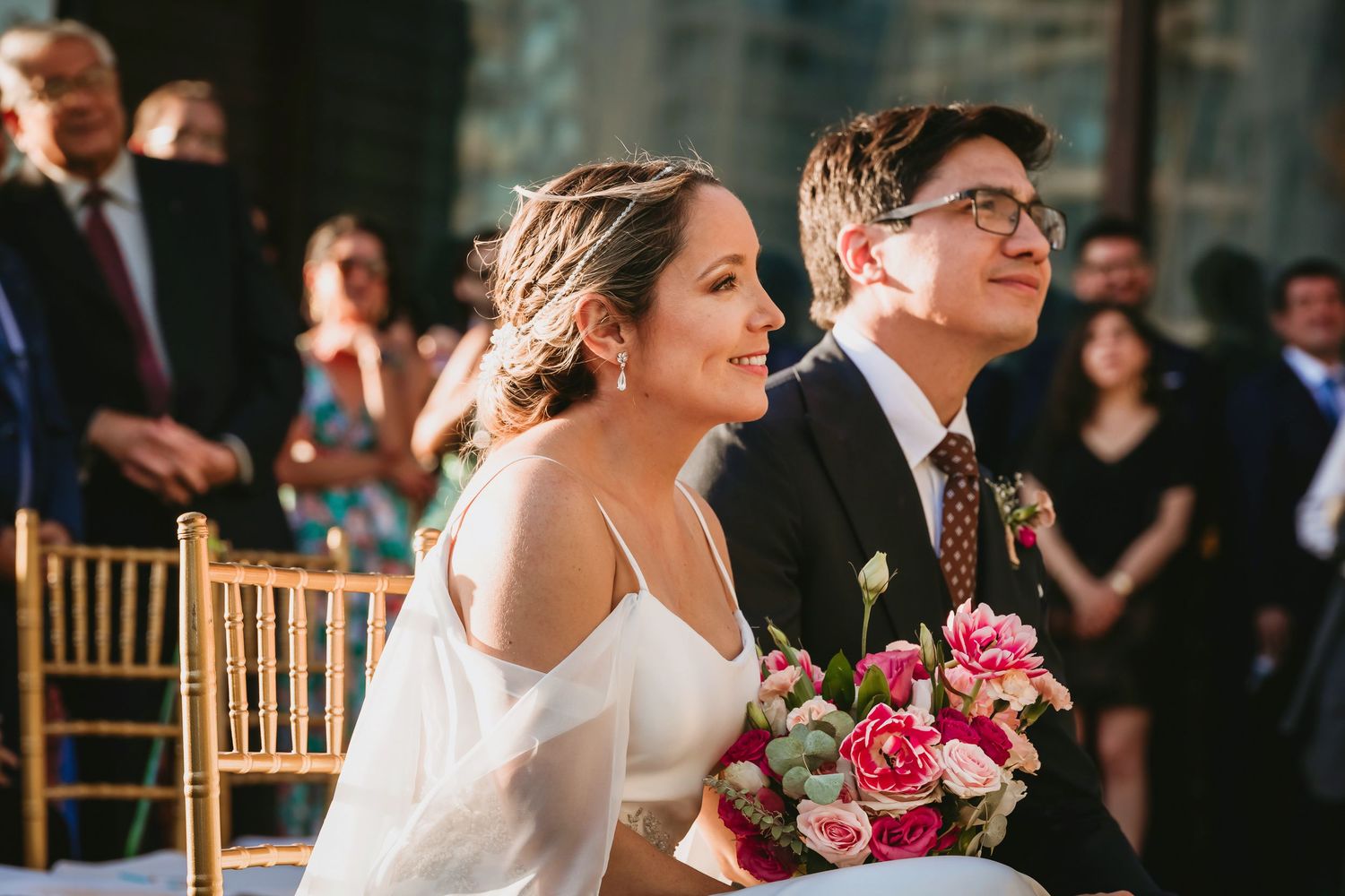 Smiling bride and groom sit together during outdoor wedding ceremony holding pink bouquet.