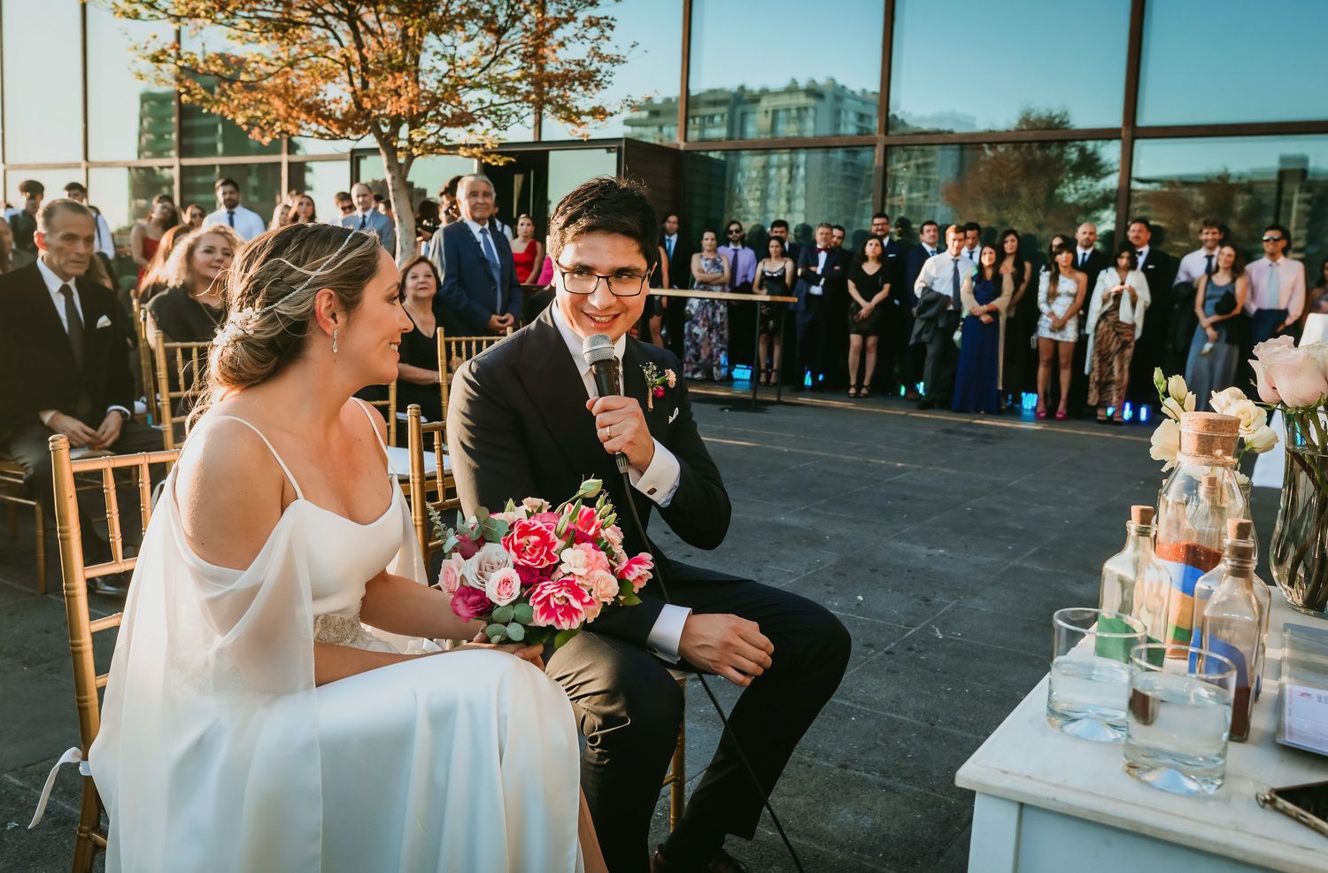 Bride and groom sit laughing during outdoor wedding ceremony with guests gathered behind them.
