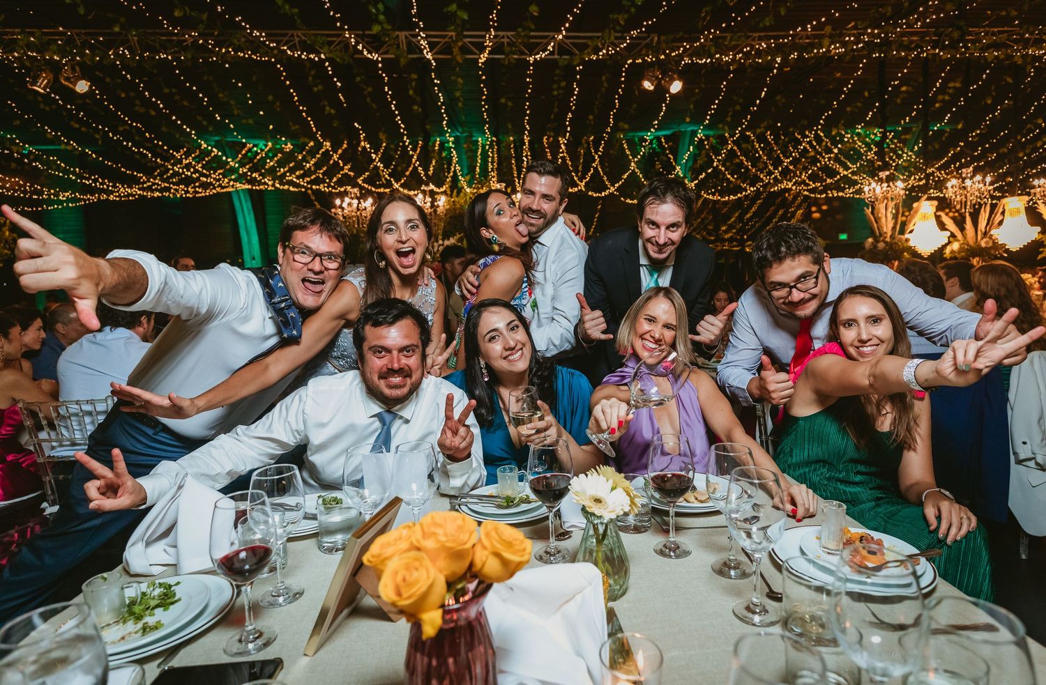 Excited group of wedding guests posing together at a decorated dinner table under twinkling lights.