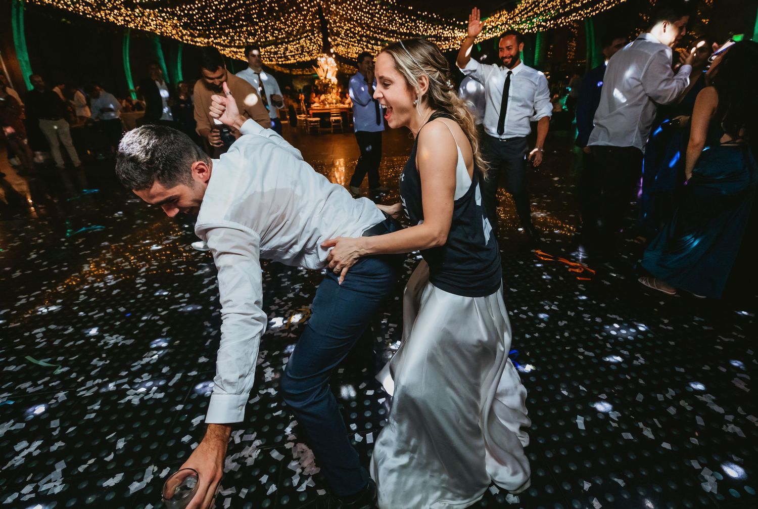 Bride in white gown dancing playfully with groom on confetti-covered dance floor.