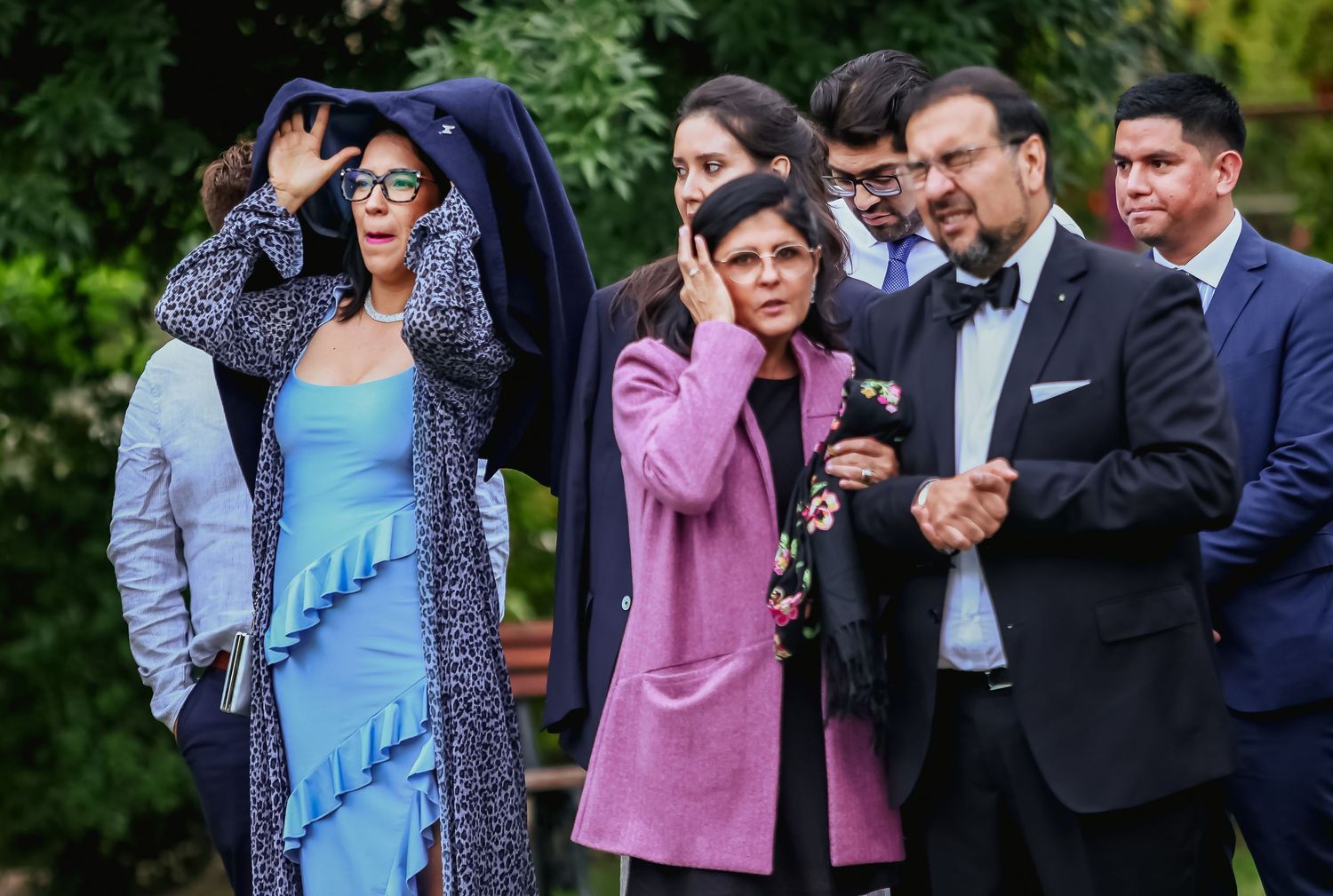 Elegantly dressed wedding guests standing outdoors, adjusting hair in windy conditions at garden event.