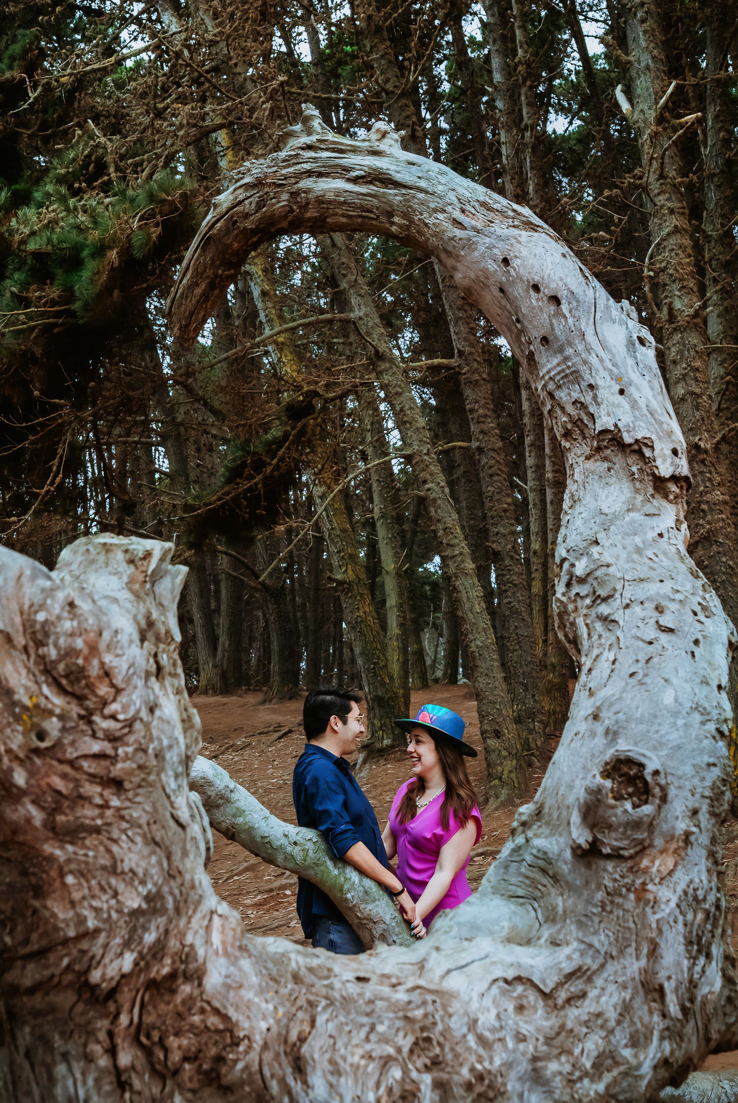 Couple holding hands framed by a twisted white tree arch in a forest, woman in pink top and blue hat.