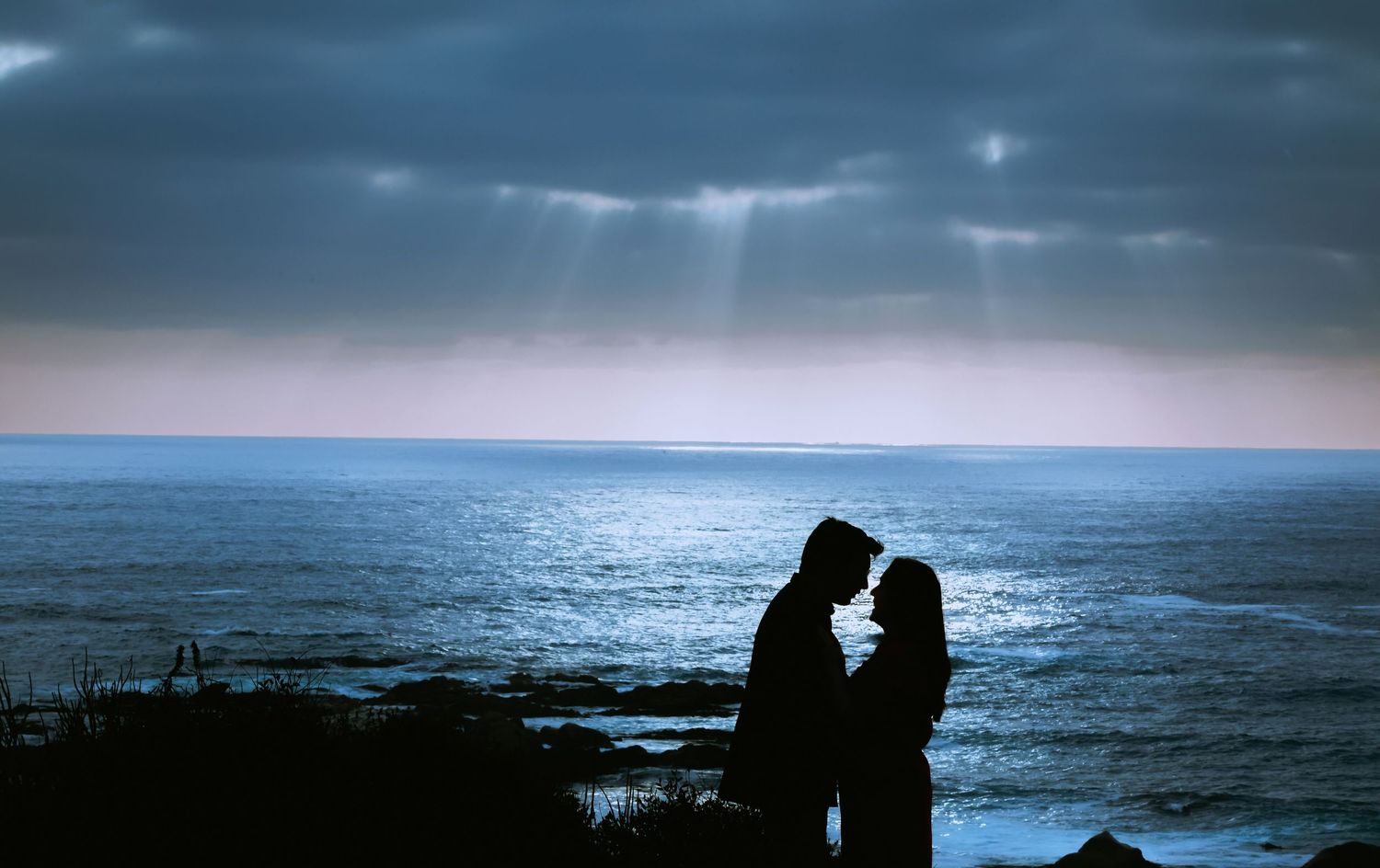 Silhouette of romantic couple facing each other by the ocean under dramatic cloudy skies with rays of light.