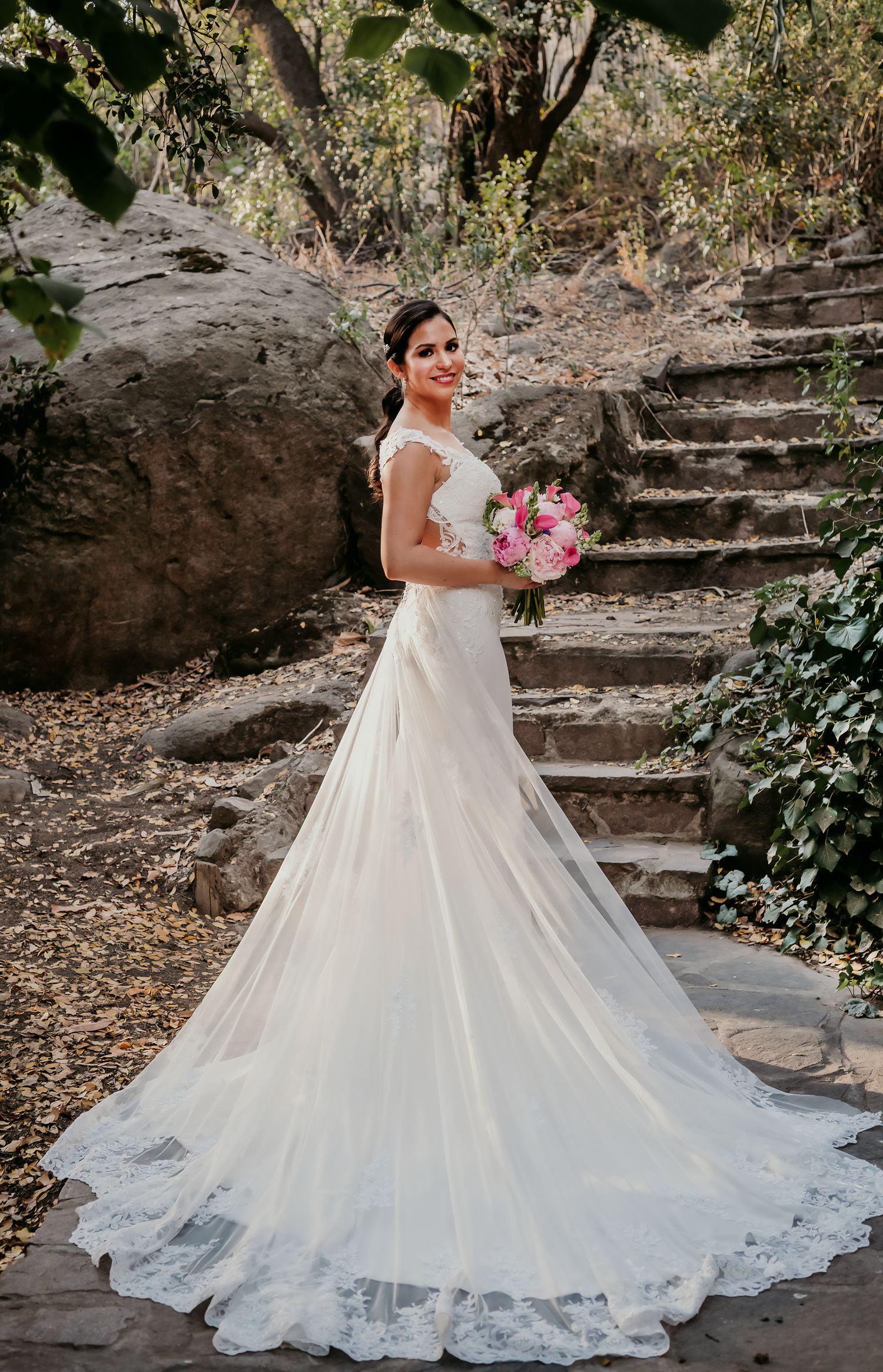 Bride in elegant white lace wedding gown with long train holds pink bouquet on stone steps in outdoor setting.