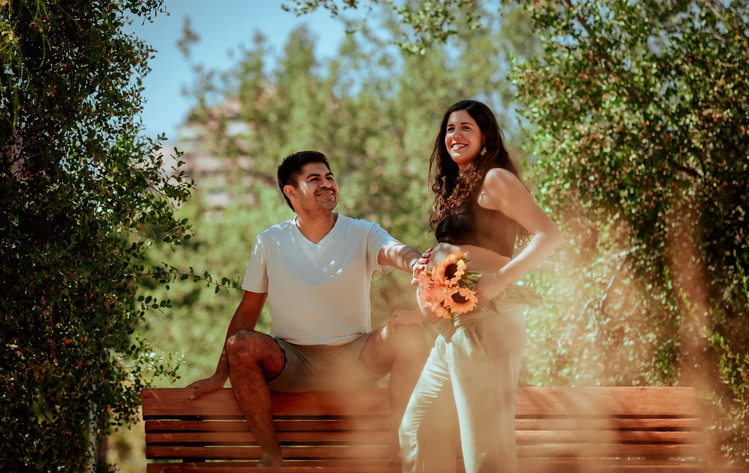 Couple sitting on a park bench surrounded by green trees, woman holding sunflowers, smiling together outdoors.