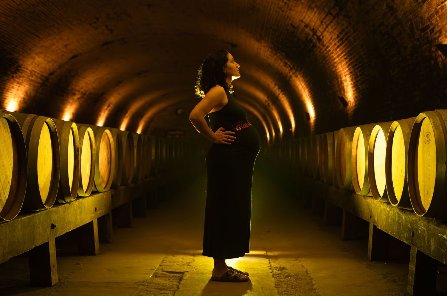 Pregnant woman in black dress stands in arched golden-lit tunnel lined with wooden wine barrels.