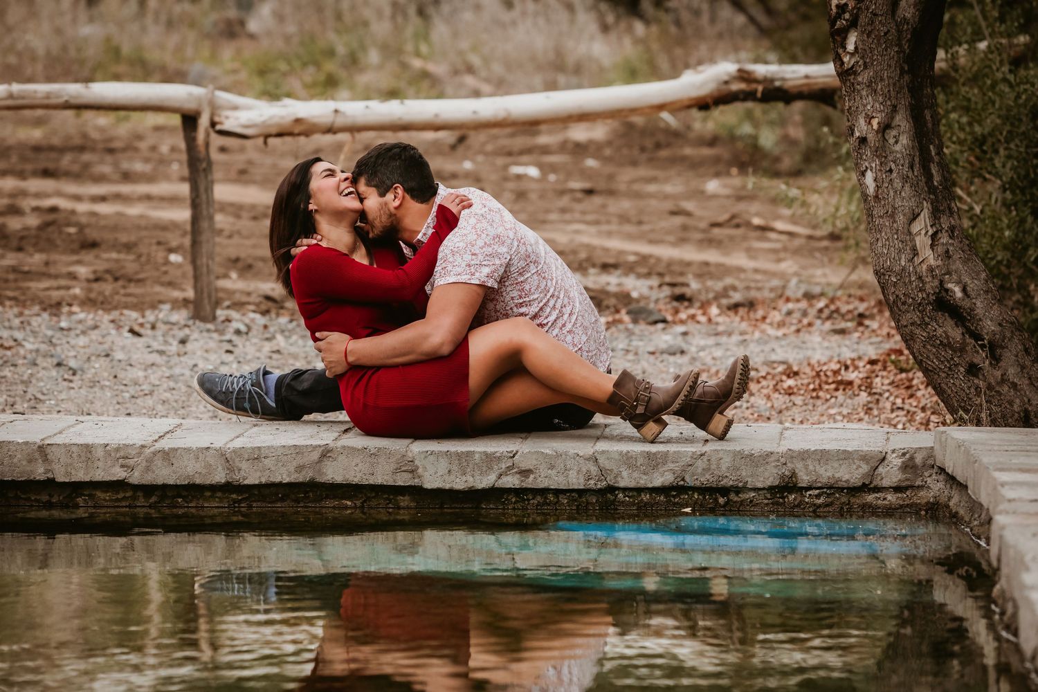 Couple kissing while sitting together beside a reflective pool in a rustic outdoor setting.