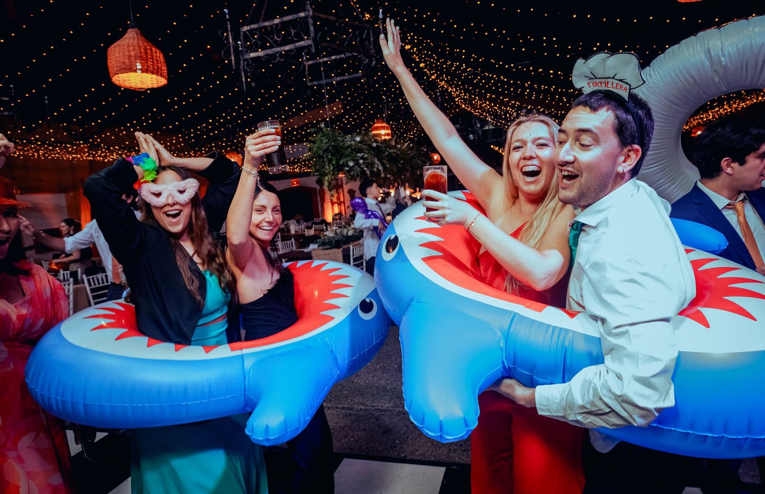 Guests dancing joyfully wearing inflatable shark costumes at an outdoor wedding reception under string lights.