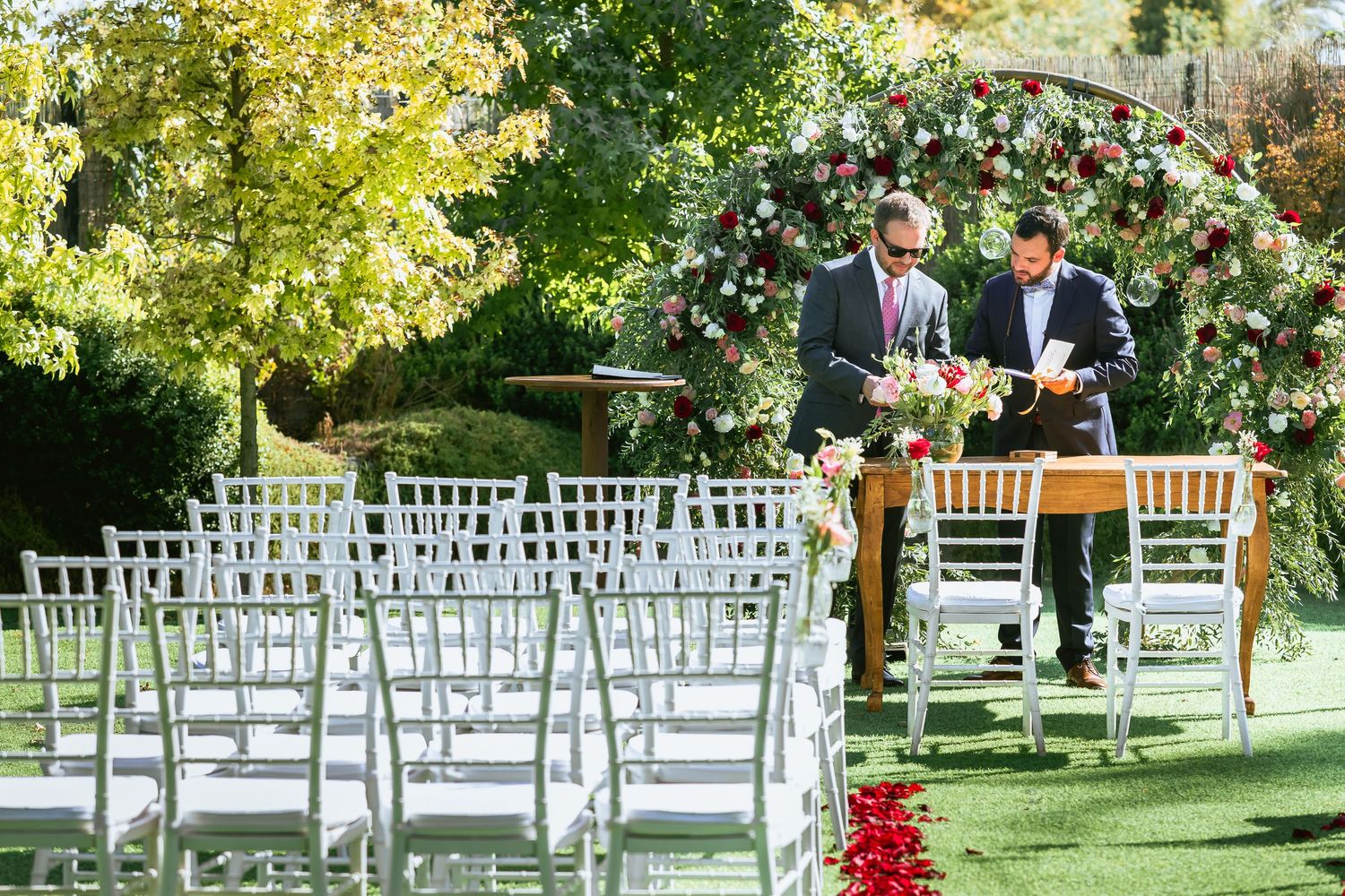 Outdoor wedding ceremony setup with white chiavari chairs and floral arch on green lawn.
