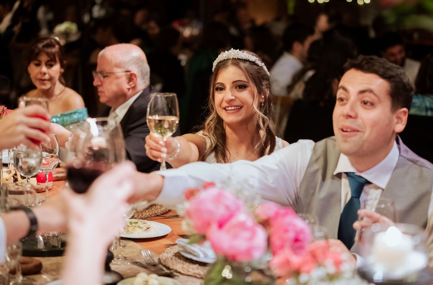 Smiling bride in tiara raises white wine glass in a toast with guests at wedding reception dinner.