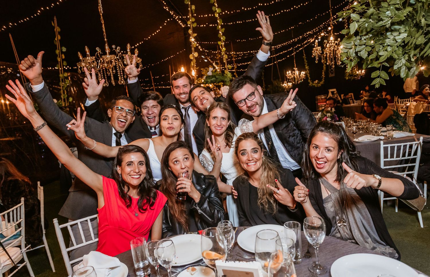 Joyful wedding guests posing together at candlelit reception table with raised hands and smiles.