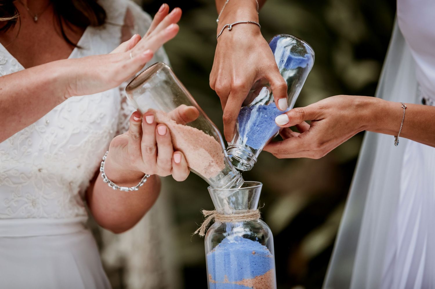 Two brides and officiant perform sand ceremony pouring colored sand into glass vase at wedding.