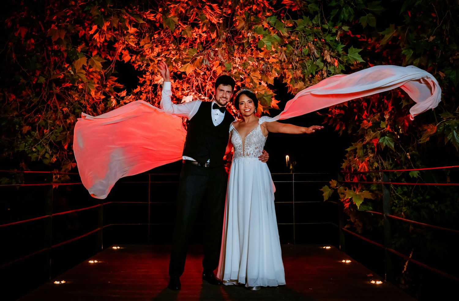 Wedding couple poses dramatically at night with bride's veil flowing in wind against red-lit foliage backdrop.