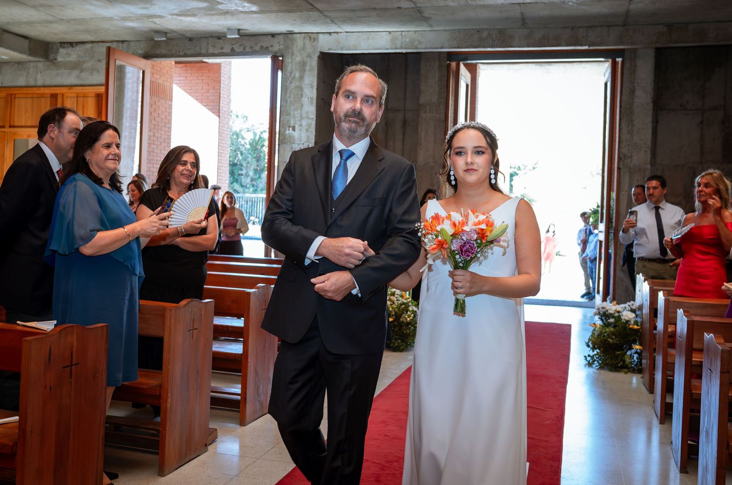 Father escorts bride in white gown down red carpet aisle in modern church.