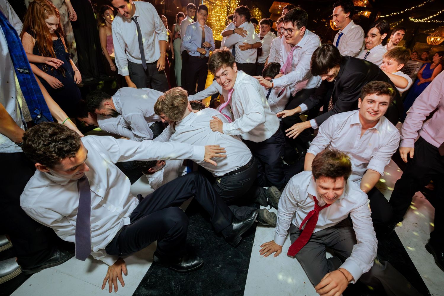Guests in formal wear tumble and pile together on white dance floor at wedding.