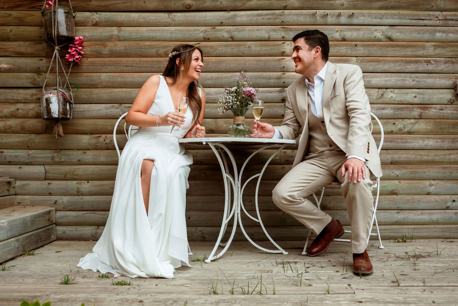 Bride and groom sit at a small white table on a rustic wooden deck, laughing together.
