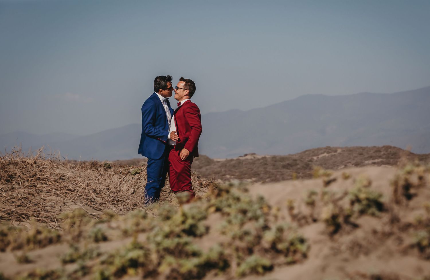 Two grooms sharing a romantic kiss amid a scenic desert landscape with mountains in the background.