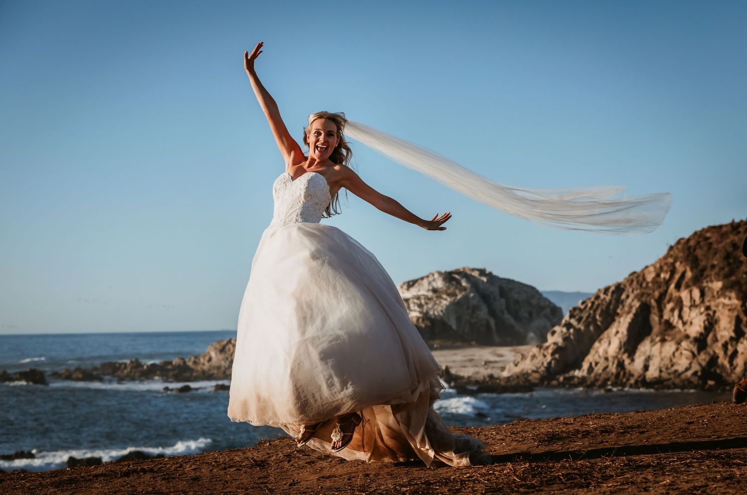 Joyful bride celebrating on rocky beach, veil soaring high against blue sky and ocean.