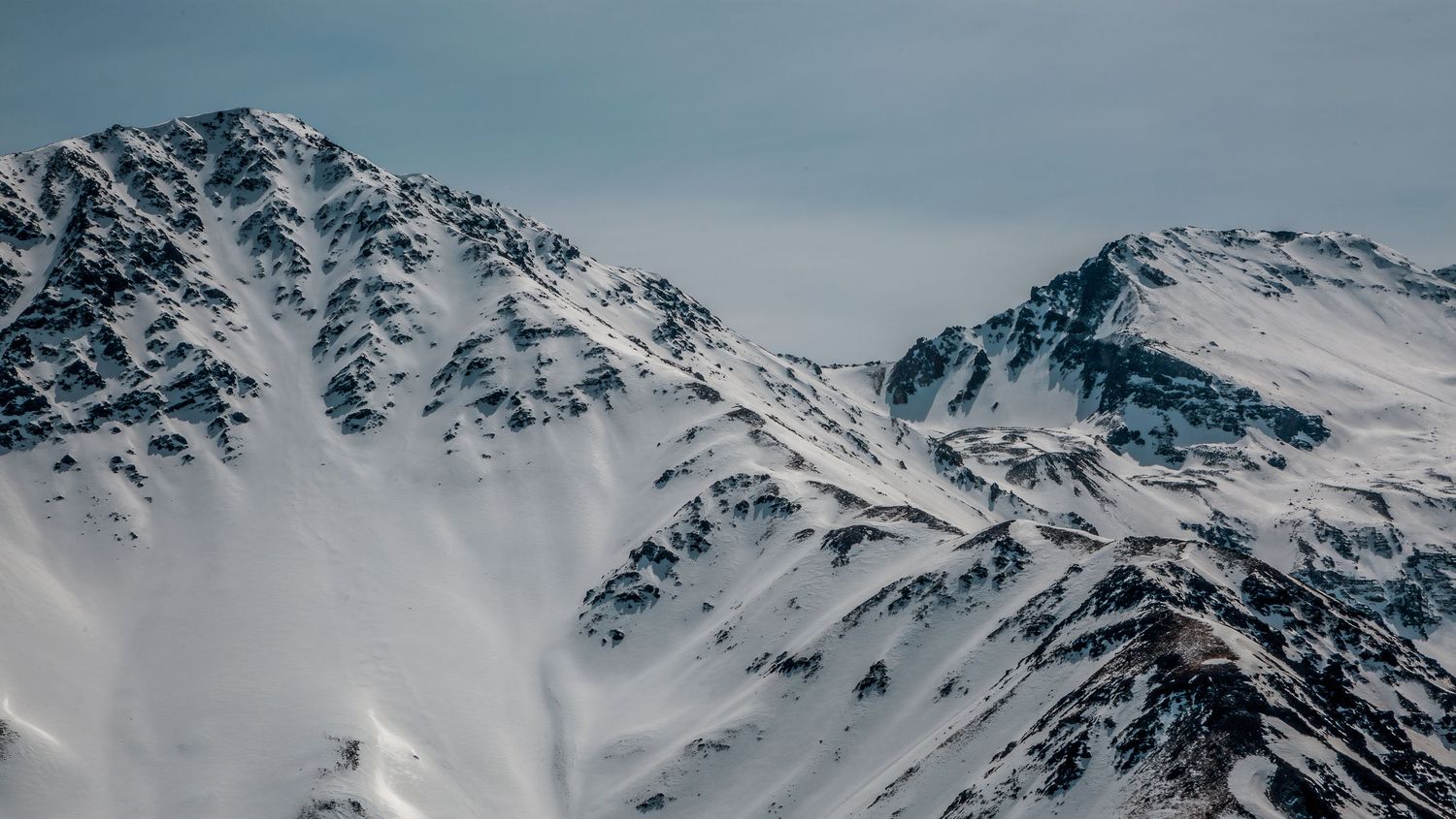 Aerial view of dramatic snow-covered mountain peaks against a pale blue winter sky.