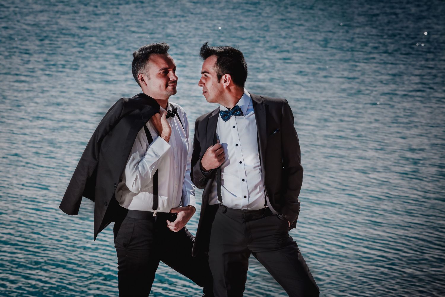 Two grooms in black suits laugh together near a shimmering blue mountain lake at dusk.