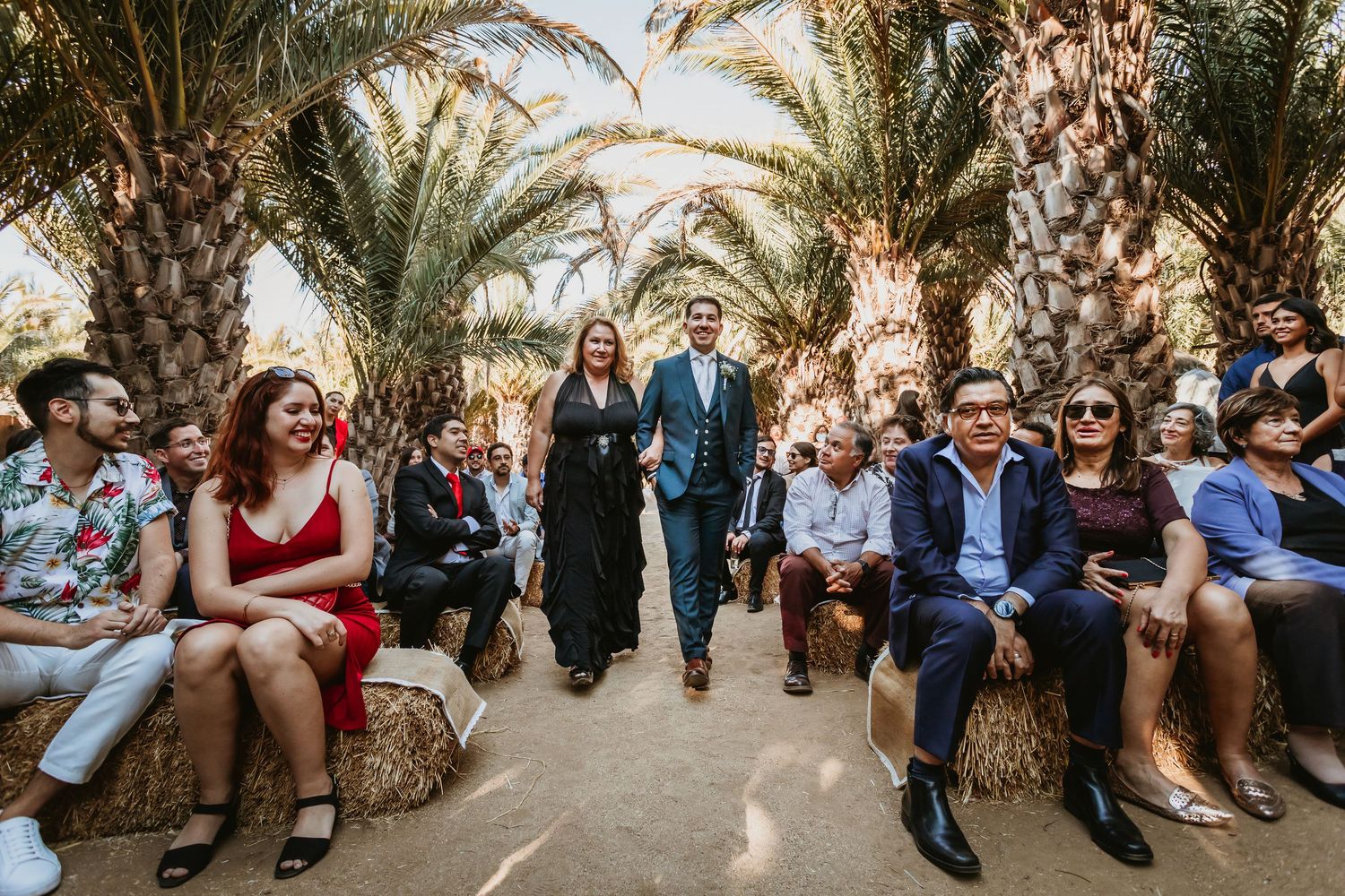 Couple walking down palm-lined wedding aisle with seated guests on hay bales outdoors.