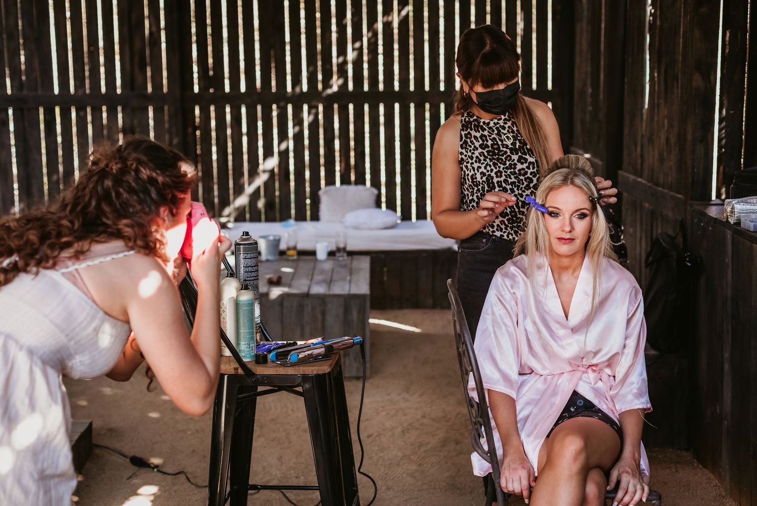 Bride in pink satin robe getting hair and makeup done in a rustic barn wedding preparation.