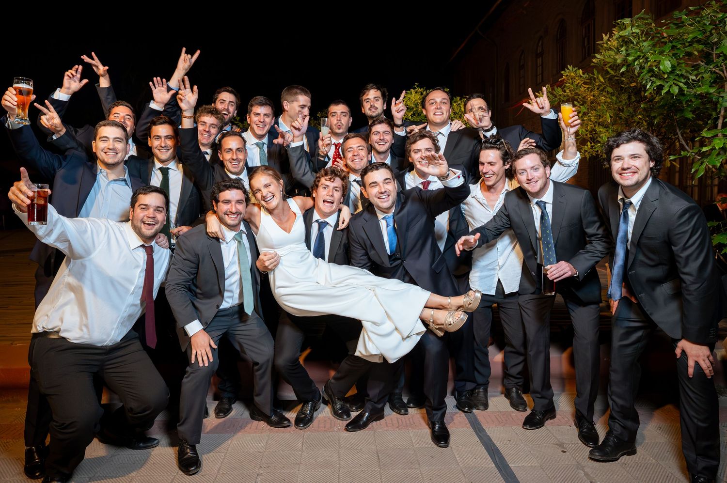 Joyful wedding party group photo with groom carrying bride surrounded by cheering guests in formal attire.