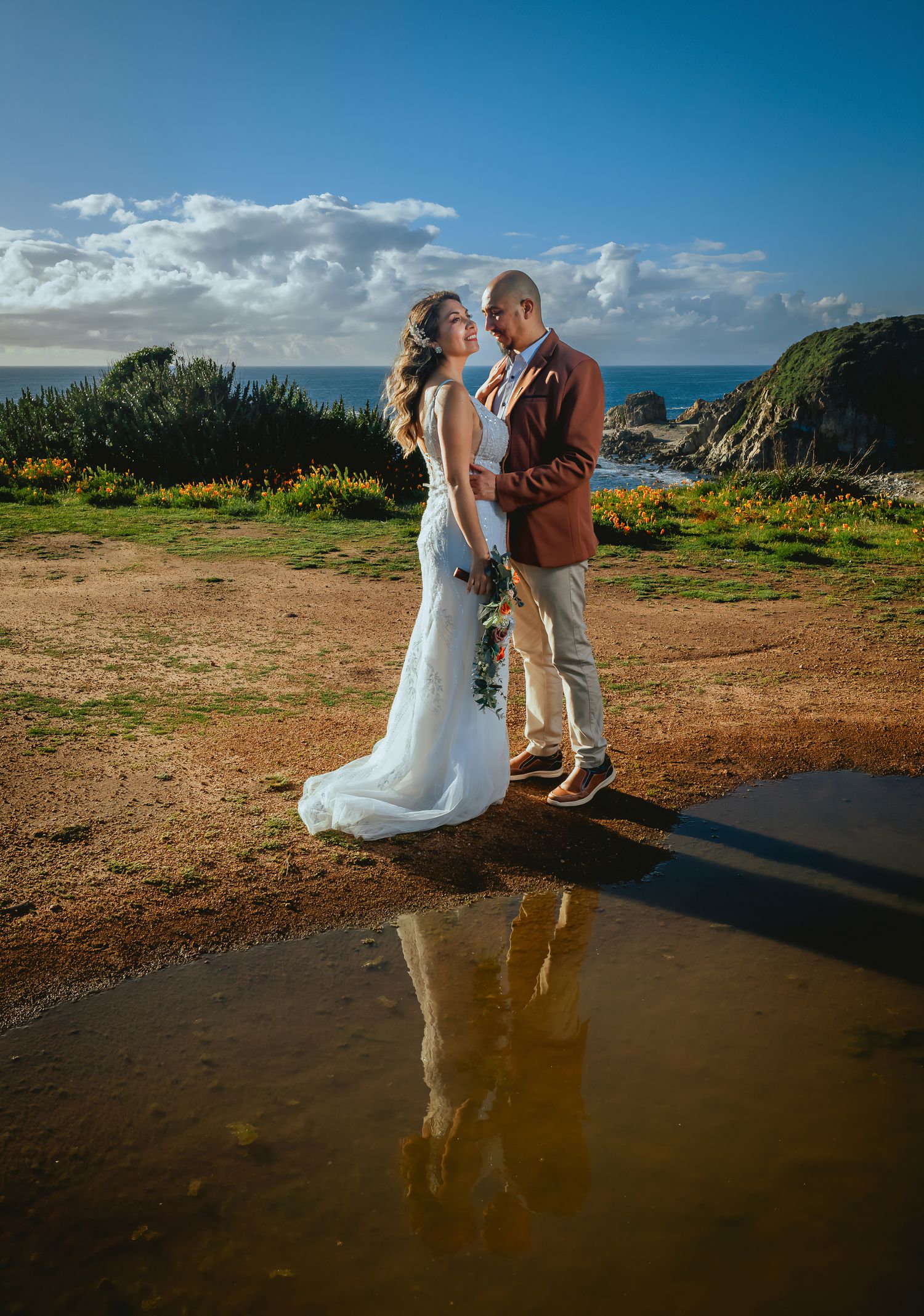 Bride in a white gown and groom in a burgundy blazer embrace on a grassy coastal cliff, their reflection glowing in a puddle at sunset.