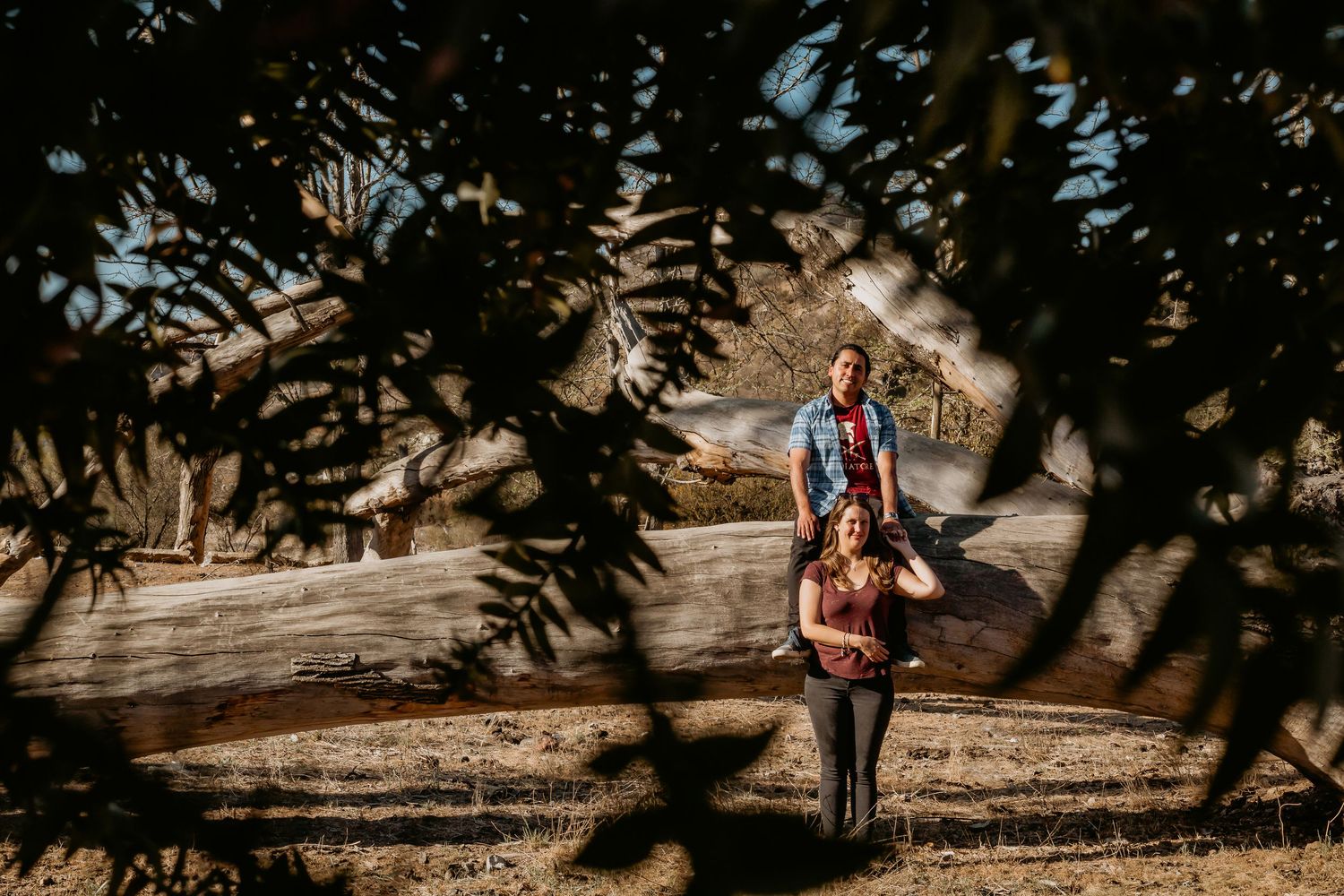 Couple posing among leafy branches with a large fallen log and dry landscape in background.
