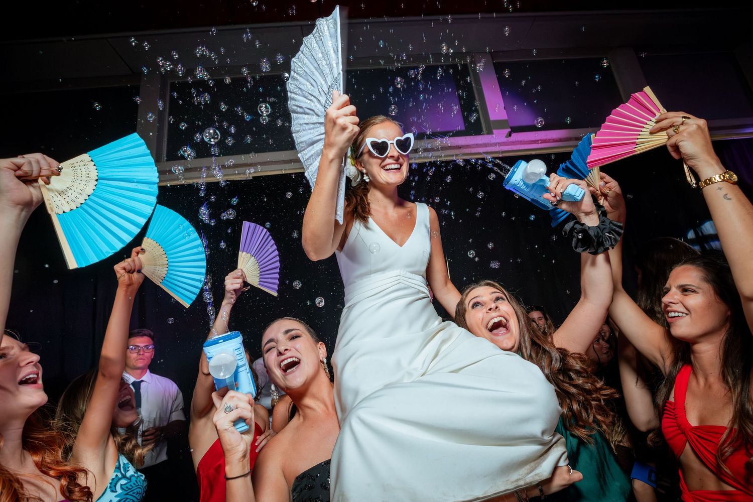 Bride lifted by guests on dance floor holding decorative fans while wearing heart-shaped sunglasses.