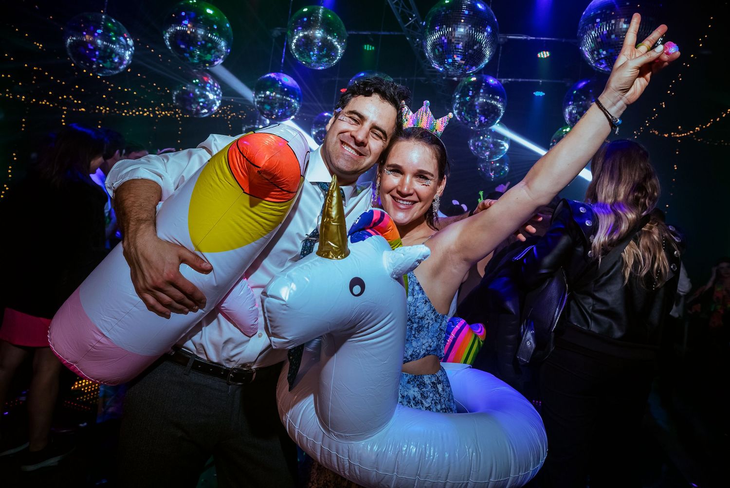 Couple posing with inflatable unicorn pool floats at a colorful nightclub party with disco balls.