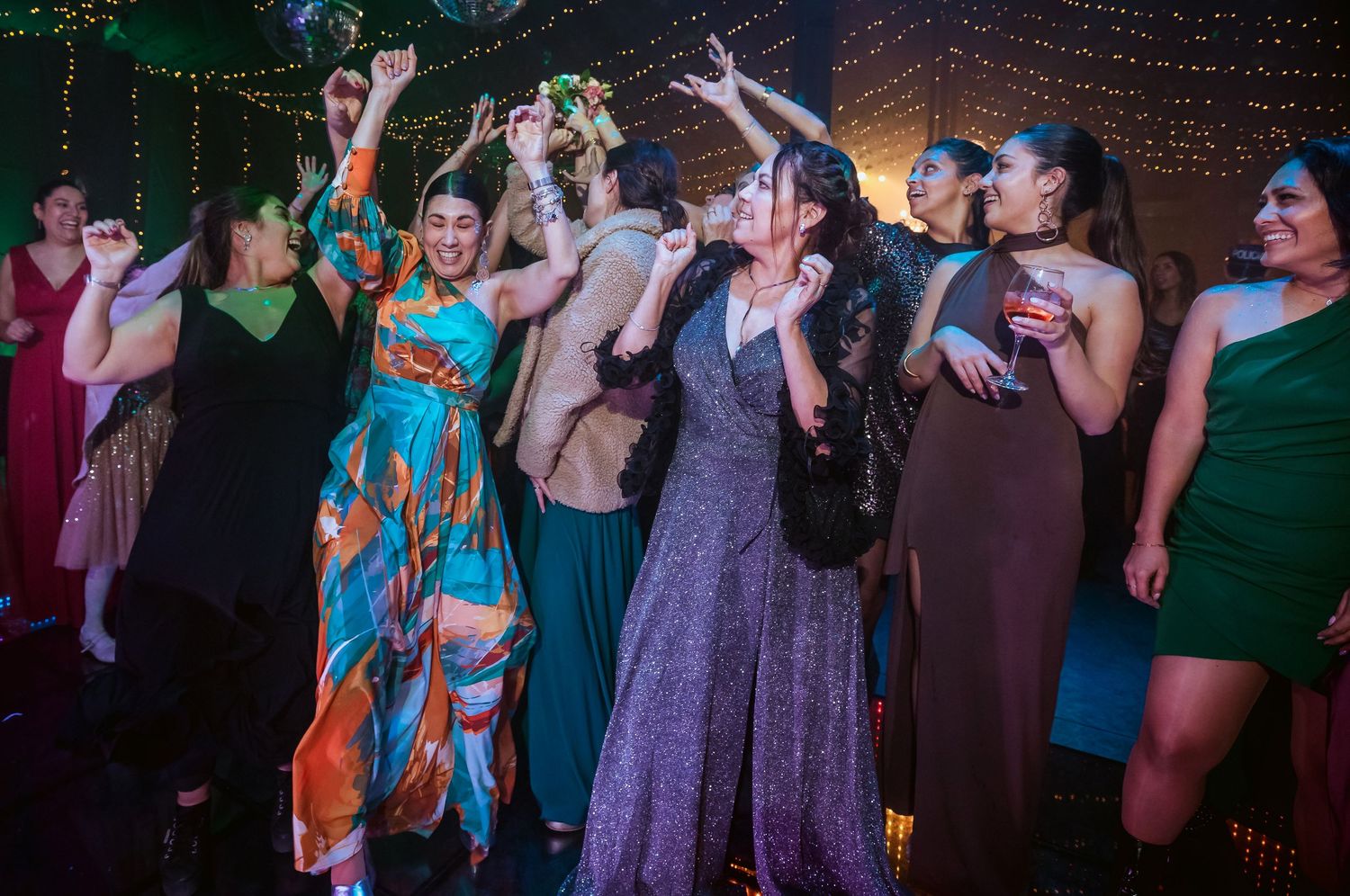 Women dancing joyfully at a festive party, wearing elegant dresses in teal, green, and purple under warm lights.