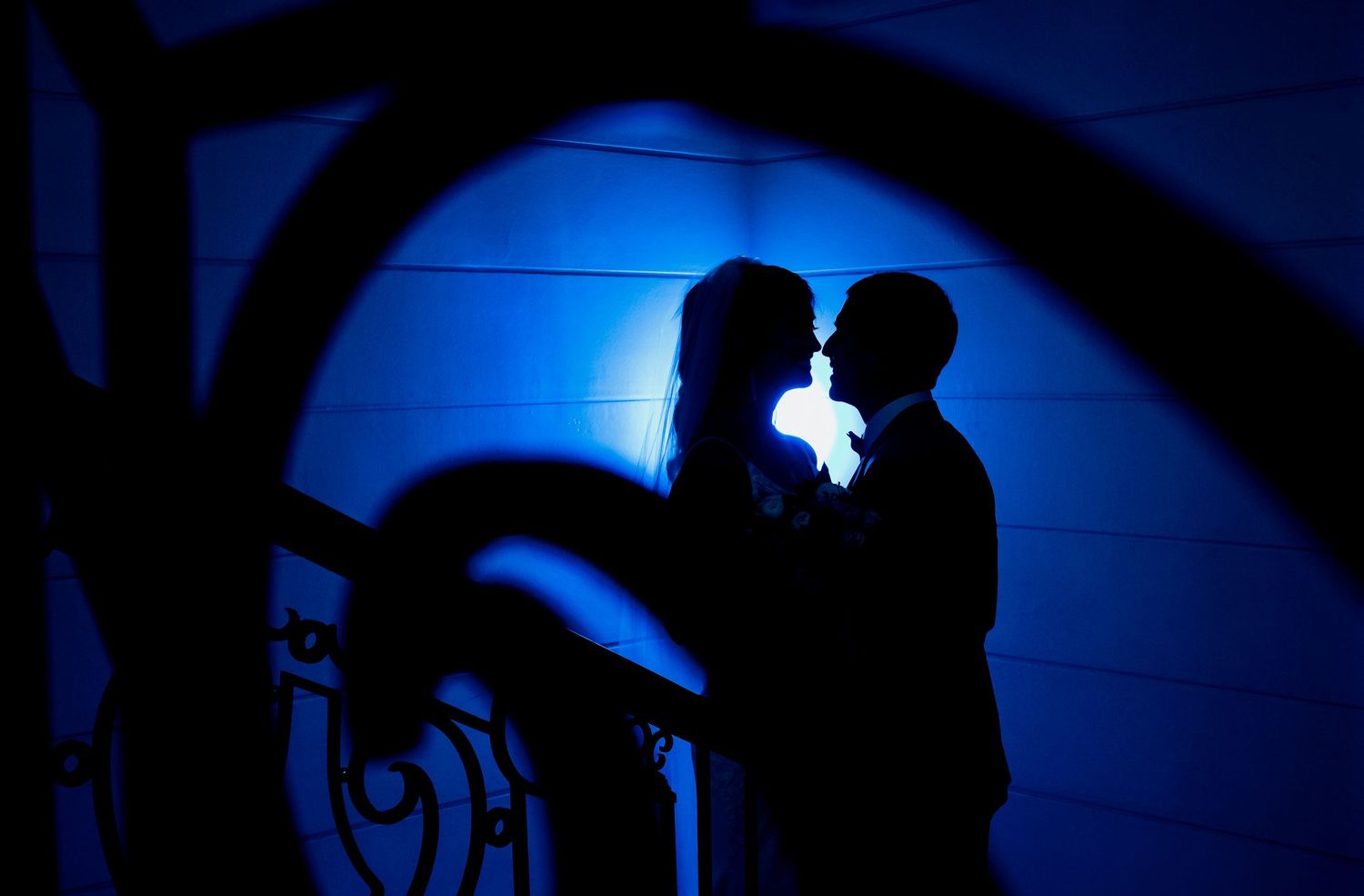 Romantic couple silhouetted against blue light, sharing a tender moment on a staircase at night.