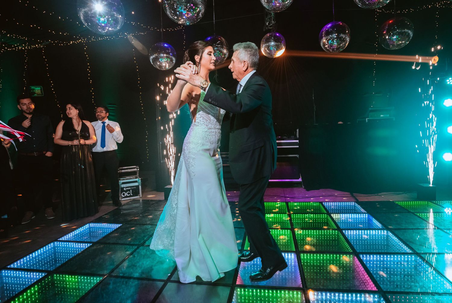 Bride and older man share first dance on colorful LED dance floor with disco balls at elegant wedding reception.