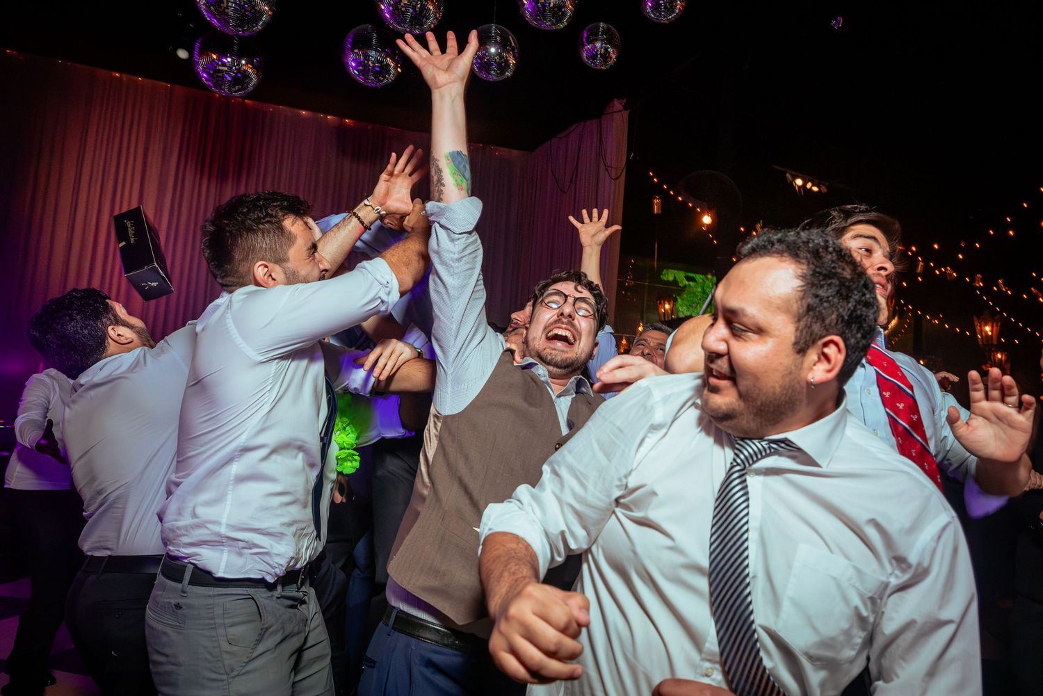 Excited wedding guests celebrate on dance floor under disco ball at reception.