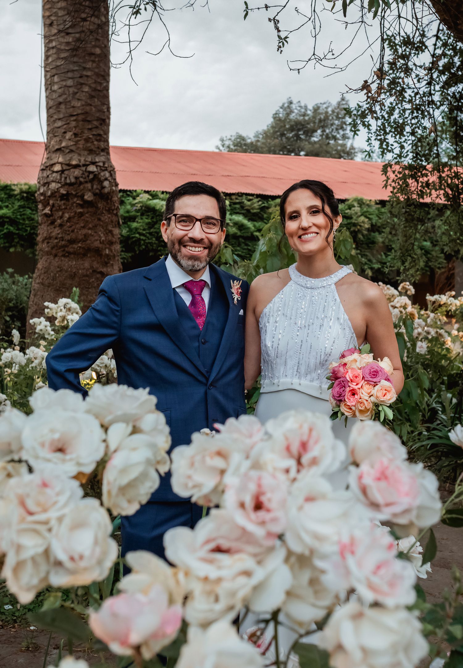 Smiling bride and groom pose together surrounded by white and pink roses at wedding.