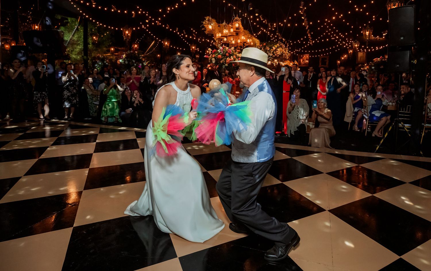 Bride and guest dance with colorful props on checkered floor surrounded by wedding crowd.