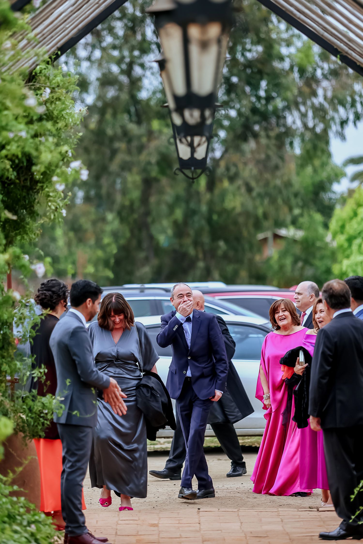 Wedding guests arriving along garden pathway with lanterns and lush green trees at outdoor venue.