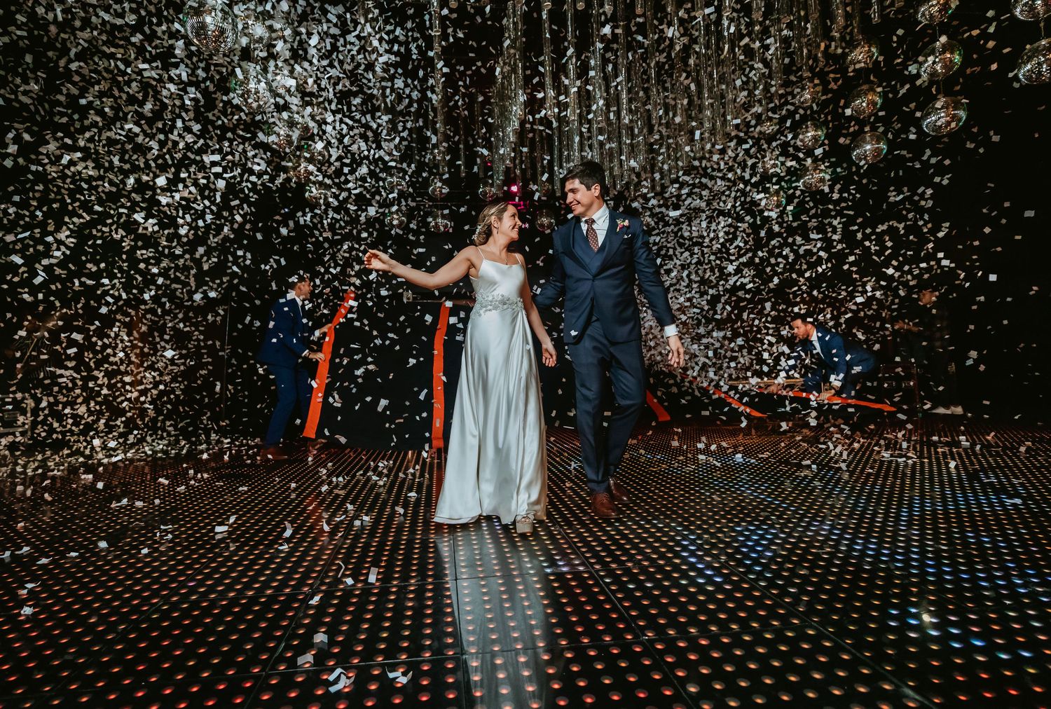 Bride and groom walking through confetti shower on sparkling dance floor at wedding.