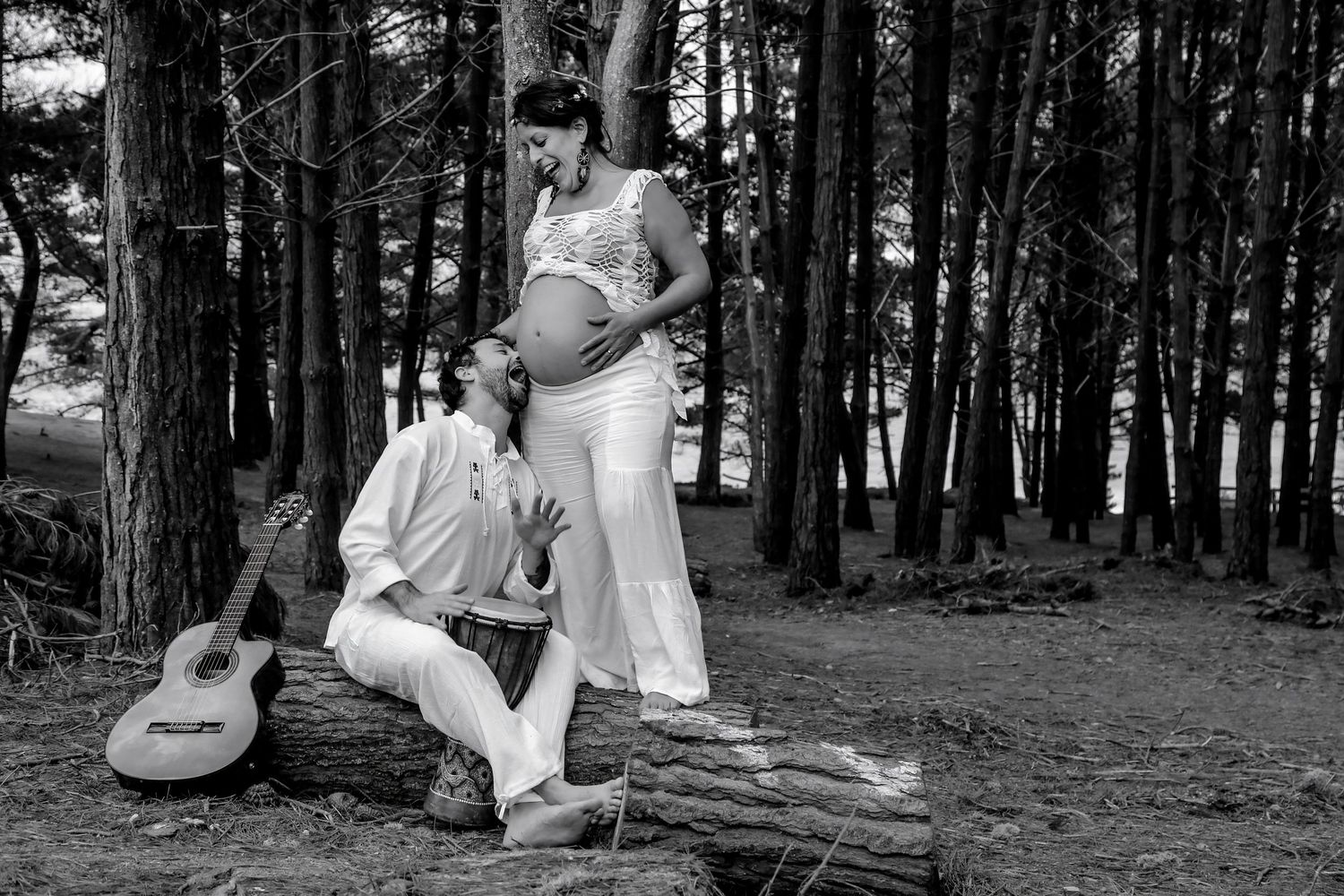 Black and white maternity photo of a couple in a forest, man sitting on a log with guitar, woman standing showing baby bump.